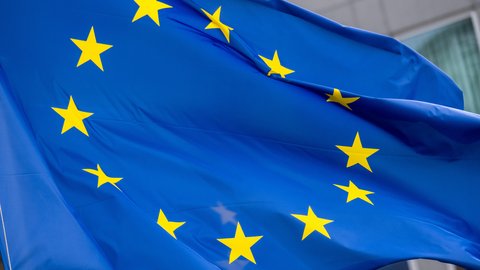 Nahaufnahme der wehenden Europaflagge; leuchtend blaues Tuch mit einem Kreis aus zwölf gelben fünfzackigen Sternen, Falten und Bewegung der Stoffbahn sichtbar, unscharfer Gebäudefronthintergrund.