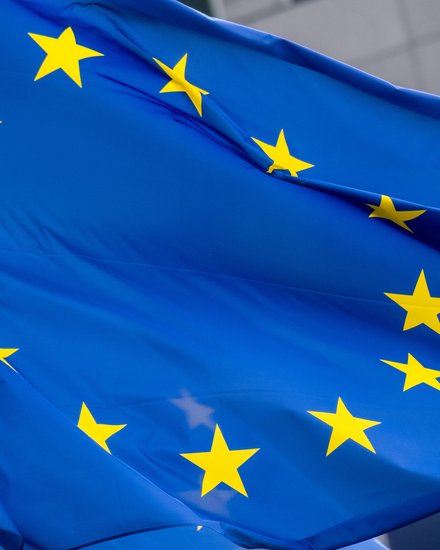 Nahaufnahme der wehenden Europaflagge; leuchtend blaues Tuch mit einem Kreis aus zwölf gelben fünfzackigen Sternen, Falten und Bewegung der Stoffbahn sichtbar, unscharfer Gebäudefronthintergrund.