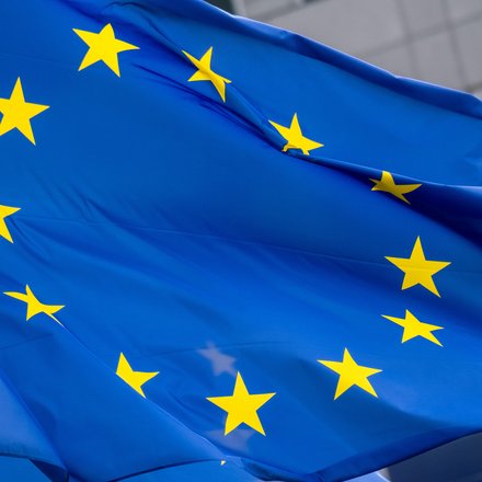 Nahaufnahme der wehenden Europaflagge; leuchtend blaues Tuch mit einem Kreis aus zwölf gelben fünfzackigen Sternen, Falten und Bewegung der Stoffbahn sichtbar, unscharfer Gebäudefronthintergrund.