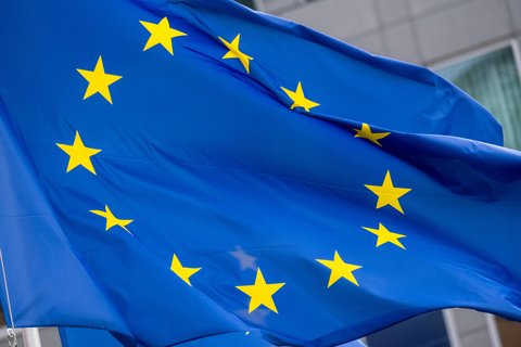Nahaufnahme der wehenden Europaflagge; leuchtend blaues Tuch mit einem Kreis aus zwölf gelben fünfzackigen Sternen, Falten und Bewegung der Stoffbahn sichtbar, unscharfer Gebäudefronthintergrund.