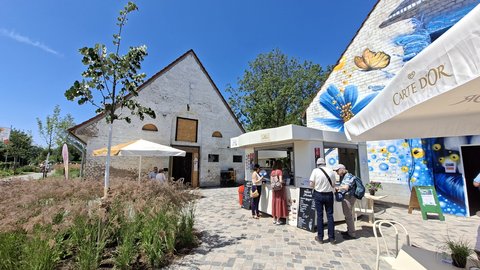 Ein Außenbereich mit modernem Café und historischem Gebäude. Ein weißes Backsteinhaus mit Satteldach steht neben zeitgenössischen weißen Pavillons mit Blumenmotiven. Mehrere Besucher stehen an den Ständen. Weiße Sonnenschirme und bepflanzte Beete prägen das Ambiente. Ein blauer Himmel ist sichtbar.