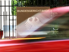 "Eingangsbereich des Bundesgerichtshofes mit braunem Schild, weißem Bundesadler-Logo und schwarzem Metallgitter. Rote Lichtstrahlen überlagern die Aufnahme. Im Hintergrund grüne Vegetation und verschwommene Gebäudeteile."