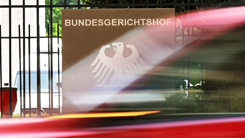"Eingangsbereich des Bundesgerichtshofes mit braunem Schild, weißem Bundesadler-Logo und schwarzem Metallgitter. Rote Lichtstrahlen überlagern die Aufnahme. Im Hintergrund grüne Vegetation und verschwommene Gebäudeteile."