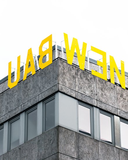 "Ausschnitt eines modernen Geschäftsgebäudes mit Eckkante. Das Gebäude hat eine grau gemusterte Fassade mit mehreren großen Fensterflächenreihen. Oben auf dem Dach prangt in großen gelben Buchstaben das Schriftzug „LADWEN". Der Himmel im Hintergrund ist bedeckt."