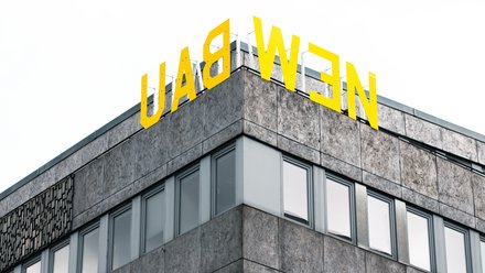 "Ausschnitt eines modernen Geschäftsgebäudes mit Eckkante. Das Gebäude hat eine grau gemusterte Fassade mit mehreren großen Fensterflächenreihen. Oben auf dem Dach prangt in großen gelben Buchstaben das Schriftzug „LADWEN". Der Himmel im Hintergrund ist bedeckt."