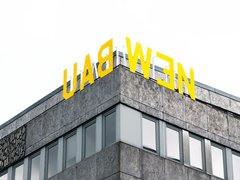 "Ausschnitt eines modernen Geschäftsgebäudes mit Eckkante. Das Gebäude hat eine grau gemusterte Fassade mit mehreren großen Fensterflächenreihen. Oben auf dem Dach prangt in großen gelben Buchstaben das Schriftzug „LADWEN". Der Himmel im Hintergrund ist bedeckt."