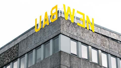 "Ausschnitt eines modernen Geschäftsgebäudes mit Eckkante. Das Gebäude hat eine grau gemusterte Fassade mit mehreren großen Fensterflächenreihen. Oben auf dem Dach prangt in großen gelben Buchstaben das Schriftzug „LADWEN". Der Himmel im Hintergrund ist bedeckt."