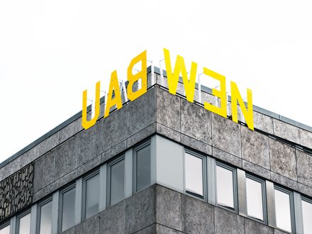 "Ausschnitt eines modernen Geschäftsgebäudes mit Eckkante. Das Gebäude hat eine grau gemusterte Fassade mit mehreren großen Fensterflächenreihen. Oben auf dem Dach prangt in großen gelben Buchstaben das Schriftzug „LADWEN". Der Himmel im Hintergrund ist bedeckt."