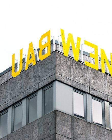 "Ausschnitt eines modernen Geschäftsgebäudes mit Eckkante. Das Gebäude hat eine grau gemusterte Fassade mit mehreren großen Fensterflächenreihen. Oben auf dem Dach prangt in großen gelben Buchstaben das Schriftzug „LADWEN". Der Himmel im Hintergrund ist bedeckt."