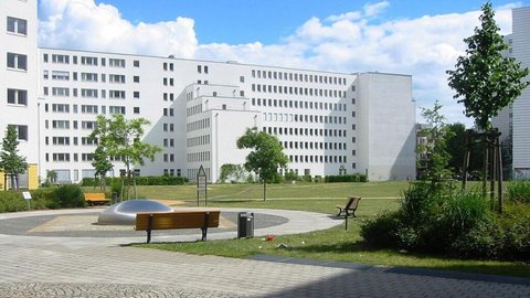 "Moderner Wohnkomplex mit mehreren weißen Mehrfamilienhäusern, Grünfläche mit Bäumen, Sitzbank mit blauem Dach und schwarzem Stuhl im Innenhofbereich."
