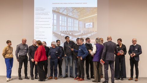 Eine Gruppe von Erwachsenen steht auf einer Bühne vor einer projizierten Präsentationsfolie, auf der ein Architekturprojekt zur Sanierung und zum Umbau eines Rathauses gezeigt wird, einige Personen halten Unterlagen in der Hand oder wenden sich einander zu, wodurch das Bild eine offizielle Preisverleihung oder Projektpräsentation dokumentiert und für Anerkennung architektonischer Qualität sowie öffentlichen Diskurs über Baukultur steht.