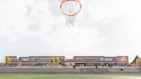 Basketballkorb im Vordergrund vor einer modernen, langgestreckten Schulgebäudefassade mit großen Fenstern und davorliegenden Stufen, auf denen Menschen sitzen und stehen