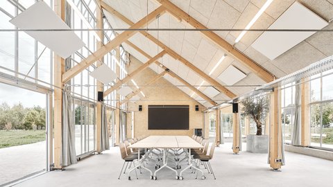 Heller Saal mit Holzfachwerk, großen Schiebefenstern und langem Konferenztisch; Akustikpaneele an der schrägen Decke.