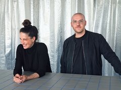 „Eine lachende Frau und ein Mann mit Brille stehen hinter einem hellgrau gefliesten Tresen. Beide tragen schwarze Kleidung; den Hintergrund bilden silbern schimmernde Vorhänge.“