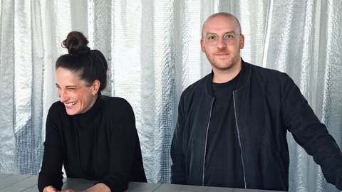 „Eine lachende Frau und ein Mann mit Brille stehen hinter einem hellgrau gefliesten Tresen. Beide tragen schwarze Kleidung; den Hintergrund bilden silbern schimmernde Vorhänge.“