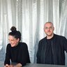 „Eine lachende Frau und ein Mann mit Brille stehen hinter einem hellgrau gefliesten Tresen. Beide tragen schwarze Kleidung; den Hintergrund bilden silbern schimmernde Vorhänge.“
