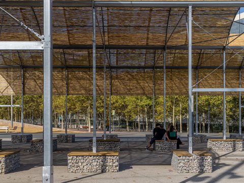 "Moderner Pavillon mit Stahlgerüst und Walmdachbedeckung aus dunklem Material. Unter dem Dach sind mehrere steinerne Sitzblöcke mit Holzauflagen angeordnet. Im Hintergrund sind grüne Bäume, ein modernes Hochhaus und weitere urban gestaltete Bereiche sichtbar. Der Boden ist gepflastert und befestigt."