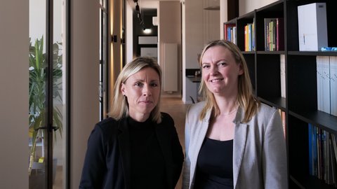 Zwei Frauen stehen in einem Büroflur vor einem Bücherregal und blicken in die Kamera. Im Hintergrund verläuft ein heller Flur mit Türen und Deckenleuchten.