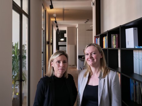Zwei Frauen stehen in einem Büroflur vor einem Bücherregal und blicken in die Kamera. Im Hintergrund verläuft ein heller Flur mit Türen und Deckenleuchten.