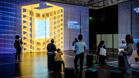 "Großer Ausstellungsraum mit bodenhoher Projektion eines gelben digitalen Gebäudemodells auf mehreren Wänden. Besucher stehen oder sitzen auf Hockern und betrachten die Darstellung. Einige fotografieren die Projektion. Text und grafische Linien ergänzen die Visualisierung und vermitteln technische Informationen über das Gebäude."
