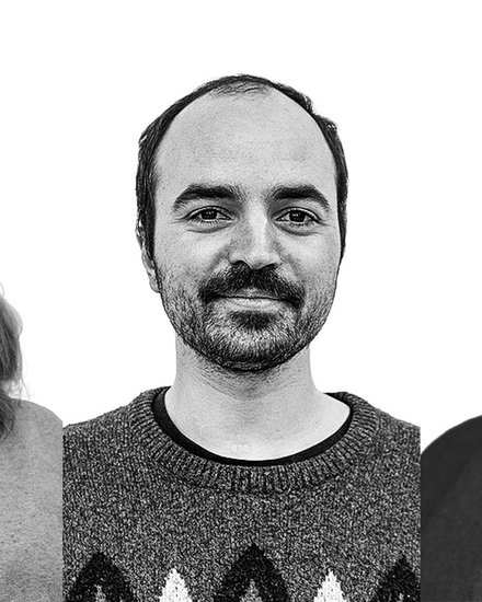 Schwarz‑Weiß‑Porträt von drei Personen nebeneinander vor weißem Hintergrund: links Person mit schulterlangem Haar und Pullover, mittig Person mit kurzem Haar und Schnurrbart im gemusterten Strickpullover, rechts Person mit kurzem Haar im Hemd; alle blicken in die Kamera und lächeln leicht.