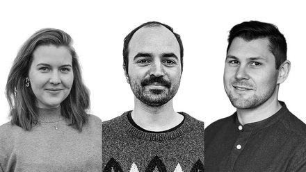 Schwarz‑Weiß‑Porträt von drei Personen nebeneinander vor weißem Hintergrund: links Person mit schulterlangem Haar und Pullover, mittig Person mit kurzem Haar und Schnurrbart im gemusterten Strickpullover, rechts Person mit kurzem Haar im Hemd; alle blicken in die Kamera und lächeln leicht.