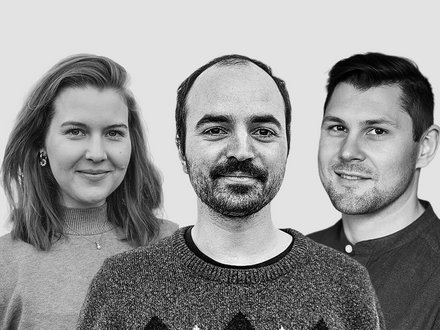 Schwarz‑Weiß‑Porträt von drei Personen nebeneinander vor weißem Hintergrund: links Person mit schulterlangem Haar und Pullover, mittig Person mit kurzem Haar und Schnurrbart im gemusterten Strickpullover, rechts Person mit kurzem Haar im Hemd; alle blicken in die Kamera und lächeln leicht.