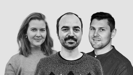 Schwarz‑Weiß‑Porträt von drei Personen nebeneinander vor weißem Hintergrund: links Person mit schulterlangem Haar und Pullover, mittig Person mit kurzem Haar und Schnurrbart im gemusterten Strickpullover, rechts Person mit kurzem Haar im Hemd; alle blicken in die Kamera und lächeln leicht.