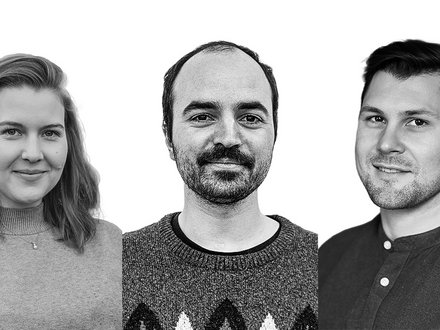 Schwarz‑Weiß‑Porträt von drei Personen nebeneinander vor weißem Hintergrund: links Person mit schulterlangem Haar und Pullover, mittig Person mit kurzem Haar und Schnurrbart im gemusterten Strickpullover, rechts Person mit kurzem Haar im Hemd; alle blicken in die Kamera und lächeln leicht.