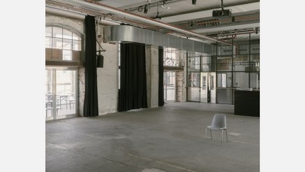 Große Industriehalle mit Betonboden, schwarzen Vorhängen und Deckenkanal; ein einzelner grauer Stuhl im leeren Raum.