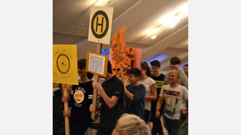 "Eine Gruppe von Jugendlichen geht durch einen Raum und hält handgemalte Schilder hoch. Ein gelbes Schild vorne zeigt ein gezeichnetes Fahrradrad mit der Aufschrift 'I want to ride my bicycle'. Ein anderes weißes Schild zeigt ein grünes 'H' im Kreis, das für eine Bushaltestelle steht. Die Jugendlichen schauen erwartungsvoll und lächeln teils."