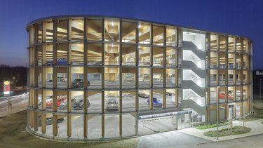 Nächtliche Außenansicht eines runden Holzparkhauses mit offener Fassadenstruktur, sichtbaren Holzstützen, beleuchteten Parkebenen und Treppenhaus.