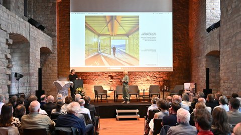 "Konferenzvortrag in einem Gewölbesaal. Auf der Bühne sitzt ein Mann im schwarzen Hemd auf der linken Seite, eine Frau in grünem Blazer steht auf der rechten Seite. Ein großer Projektorschirm zeigt eine Architektur-Visualization eines gelb beleuchteten modernes Pavillons mit Panoramafenster. Das Publikum sitzt davor im Auditorium. Die Backsteinwände sind orange beleuchtet."