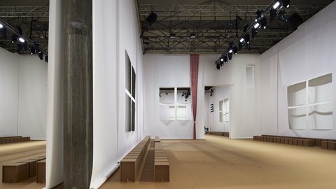 "Leerer, heller Ausstellungsraum mit hohen weißen Wänden, großen geometrischen Öffnungen und einer einzelnen karierten Stoffbahn. Holzbänke stehen entlang der Wände. Der Raum wirkt reduziert und architektonisch klar gestaltet."