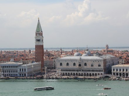 Panoramablick auf Venedig am Wasser: Links ragt der rote Campanile von San Marco mit grünem Spitzdach auf, daneben der helle Dogenpalast mit mehreren Kuppeln. Am Ufer drängen sich Menschen; auf dem türkisgrünen Wasser fahren Boote. Dahinter erstrecken sich dicht gedrängte, orange Ziegeldächer bis zum Horizont unter leicht bewölktem Himmel.