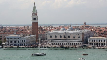 Panoramablick auf Venedig am Wasser: Links ragt der rote Campanile von San Marco mit grünem Spitzdach auf, daneben der helle Dogenpalast mit mehreren Kuppeln. Am Ufer drängen sich Menschen; auf dem türkisgrünen Wasser fahren Boote. Dahinter erstrecken sich dicht gedrängte, orange Ziegeldächer bis zum Horizont unter leicht bewölktem Himmel.
