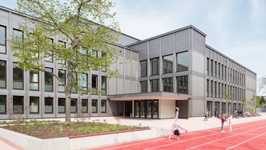 Moderner Schulbau mit Holzfassade, davor ein Schulhof mit roter Laufbahn und spielenden Kindern.