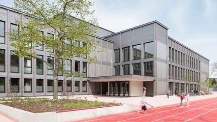 Moderner Schulbau mit Holzfassade, davor ein Schulhof mit roter Laufbahn und spielenden Kindern.