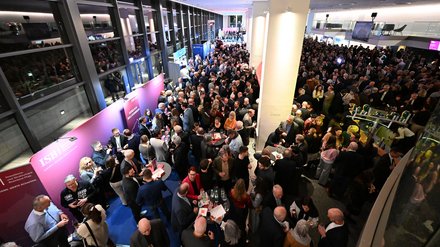 Belebtes Foyer beim Empfang: dichtes Gedränge an Stehtischen, Messestände entlang der Glasfassade; Gäste im Gespräch und beim Netzwerken.