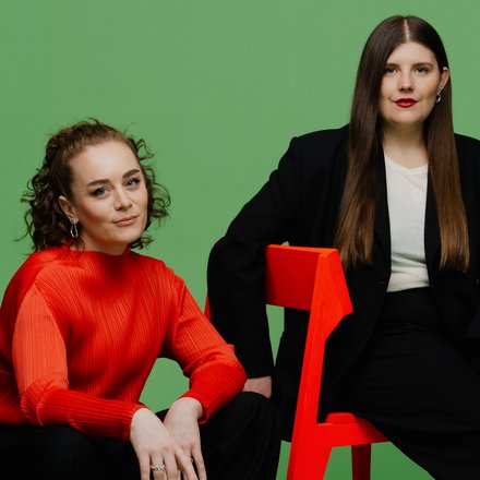  Porträt von zwei Frauen (Katharina Benjamin und Angelika Hinterbrandner) in einem Fotostudio mit grünem Hintergrund. Die linke Frau sitzt in einem leuchtend orangefarbenen Oberteil auf einem dunklen Sitzsack, die rechte Frau trägt einen schwarzen Oversize-Anzug und sitzt auf einem knallroten Stuhl. Beide blicken in die Kamera.