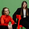  Porträt von zwei Frauen (Katharina Benjamin und Angelika Hinterbrandner) in einem Fotostudio mit grünem Hintergrund. Die linke Frau sitzt in einem leuchtend orangefarbenen Oberteil auf einem dunklen Sitzsack, die rechte Frau trägt einen schwarzen Oversize-Anzug und sitzt auf einem knallroten Stuhl. Beide blicken in die Kamera.
