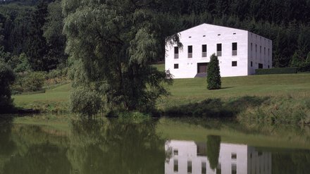 "Weißes quadratisches Betongebäude mit flachem Dach in ländlicher Waldlandschaft. Große rechteckige Fensteröffnungen auf der sichtbaren Seite. Umgeben von Wiesen und dichten grünen Wald. Klarer Himmel mit Wolken."