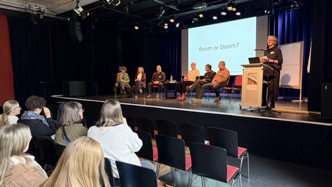 Vortragssaal mit Panel: fünf Sitzende auf der Bühne, rechts eine Person am Rednerpult. Auf der Leinwand steht „Boom or Doom?“, Publikum im Vordergrund.