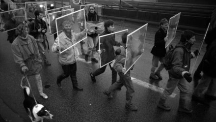 Schwarzweißaufnahme einer Demonstration aus erhöhter Perspektive: mehrere Menschen marschieren auf nasser Straße und tragen rechteckige, durchsichtige Schilder; vorne läuft ein Hund an der Leine.