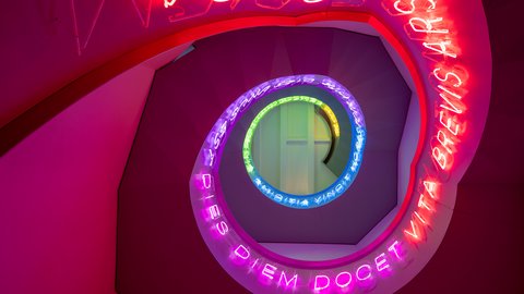 Bunte, spiralförmige Neonlichtinstallation mit lateinischem Text an einer Treppenhausdecke