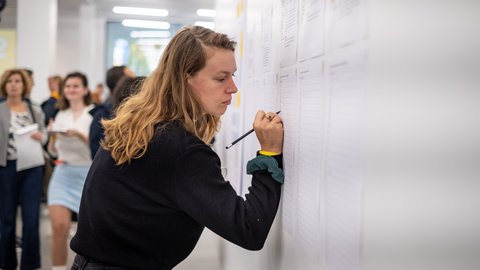 Copilot said:  Person schreibt Beiträge auf Moderationsbögen an einer Pinnwand, weitere Teilnehmende im Hintergrund.
