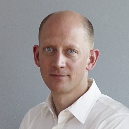 Porträtfoto einer Person in weißem Hemd vor grauer Wand, Oberkörper, neutraler Blick.