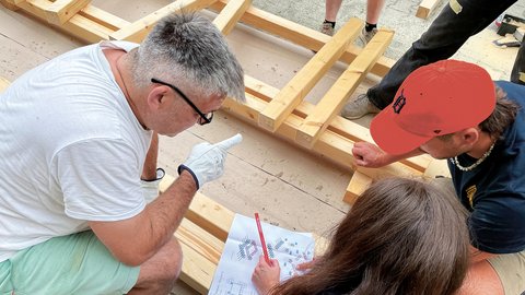 Drei Personen sitzen auf dem Boden und besprechen einen Bauplan für eine Holzkonstruktion, deren Balken neben ihnen liegen. Ein Mann mit Arbeitshandschuhen zeigt mit dem Finger auf den Bauplan."