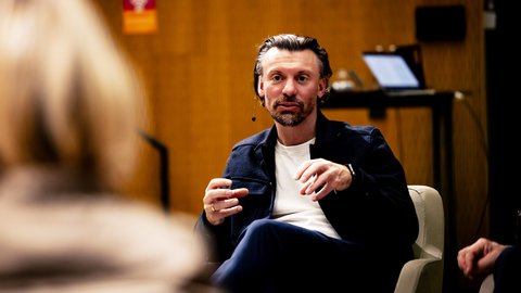  Person mit Headset‑Mikrofon spricht gestikulierend in Gesprächsrunde; unscharfe Zuhörende im Vordergrund.