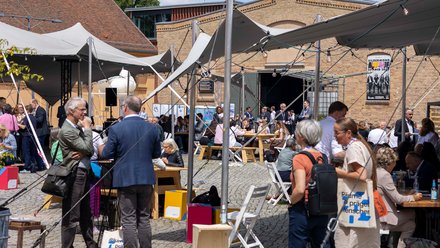 Eine Fotografie zeigt einen belebten Innenhof mit temporären Zeltdächern, Holzbänken und Tischen, zahlreiche Menschen sitzen, stehen oder kommen miteinander ins Gespräch, während historische Backsteinbauten den Rahmen bilden, wodurch das Bild eine offene Veranstaltungsatmosphäre vermittelt und für Austausch, Begegnung und Baukultur als gesellschaftliches Ereignis steht.